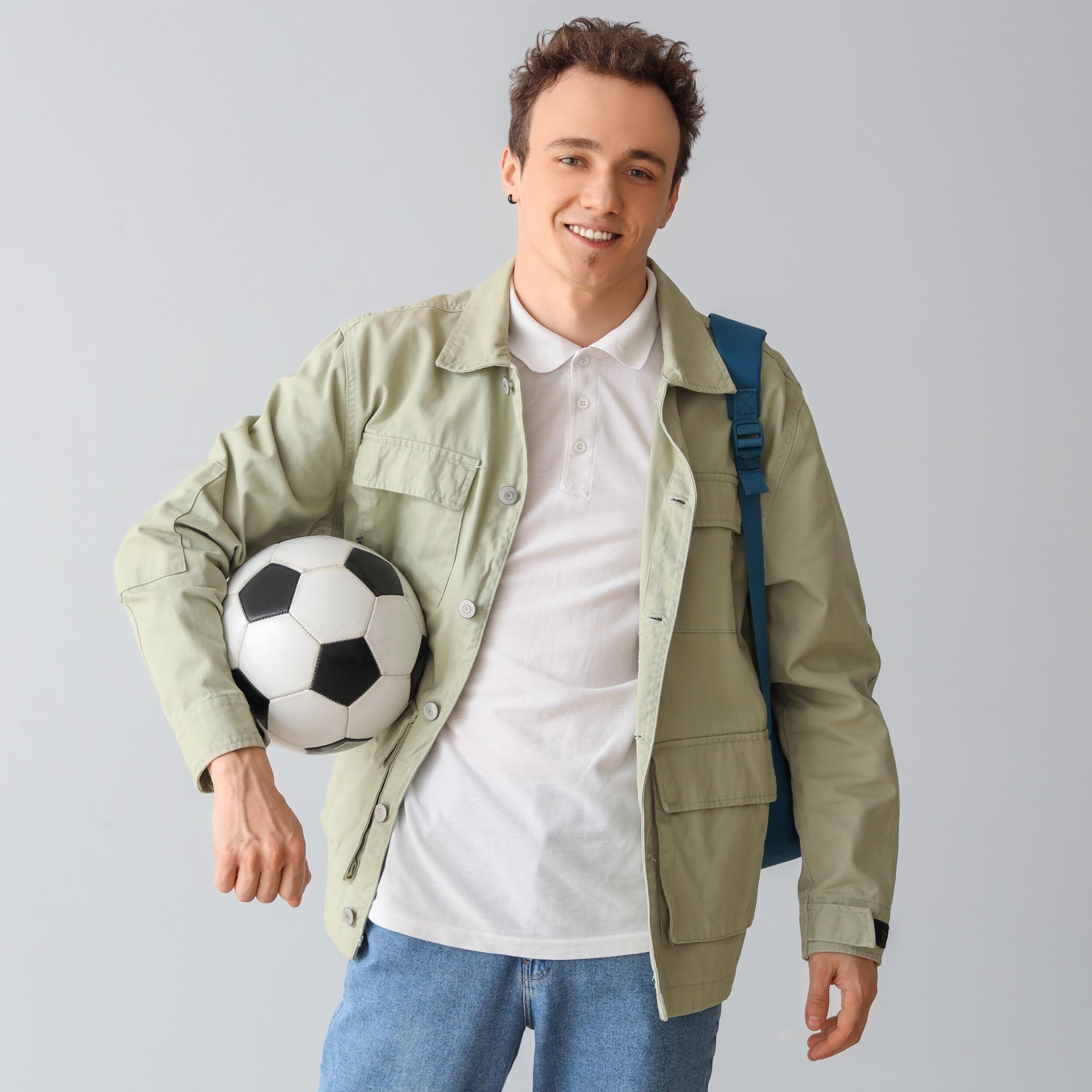 Male student with backpack and soccer ball on grey background.